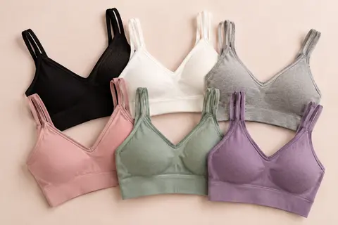 Six bralettes camisoles aux coloris noir, blanc, gris, rose, vert et violet, pliées sur un fond crème.